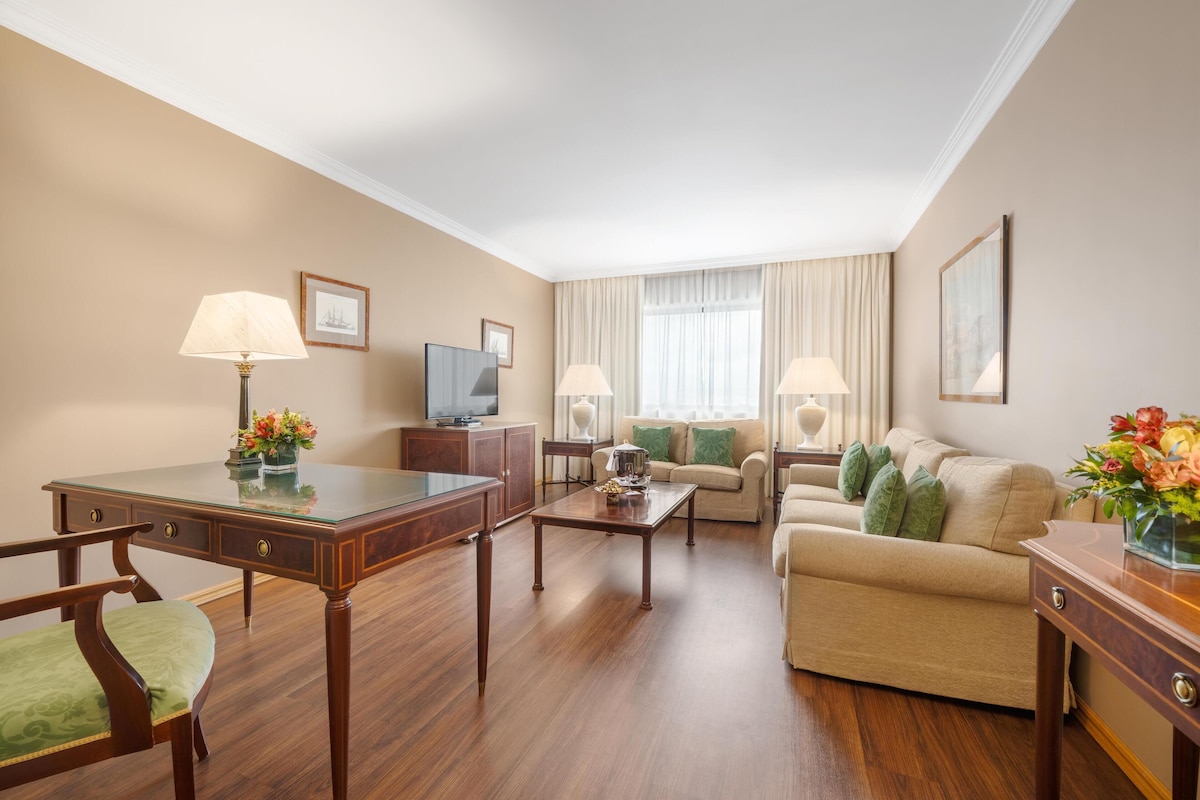 A hotel room with a wooden desk, two chairs, a sofa, a television, and two lamps.