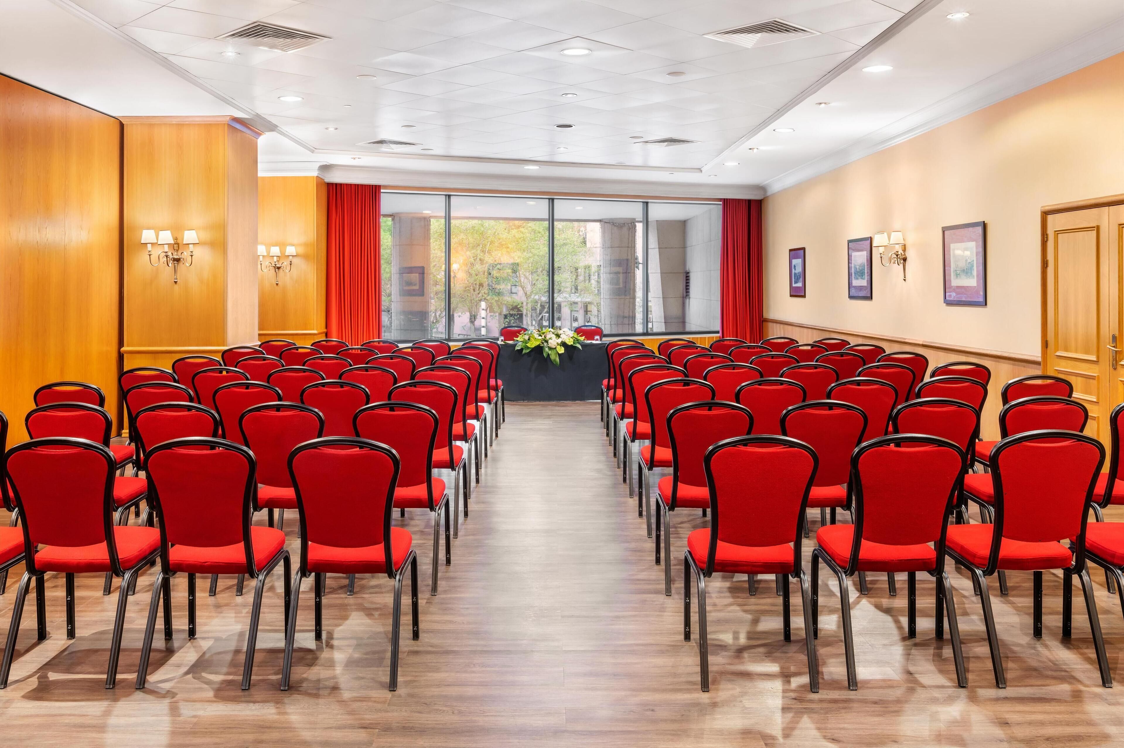 Un salón de conferencias con hileras de sillas rojas, ventanas amplias y un panel de pared de madera.
