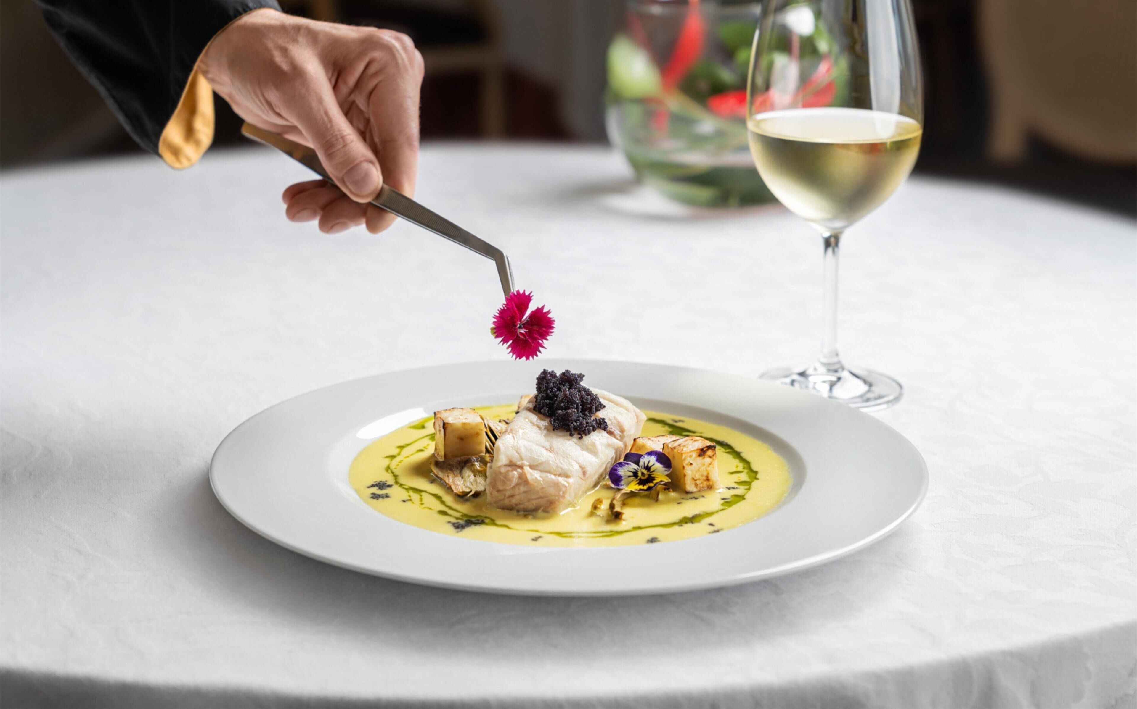 Une personne utilise une fourchette pour prendre un morceau de nourriture dans une assiette blanche. On aperçoit en arrière-plan un verre de vin blanc et un vase de fleurs.