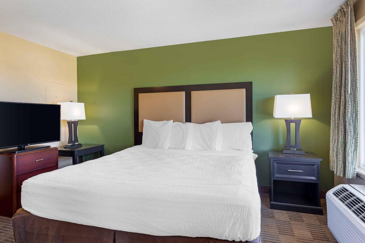 A hotel room with a large bed, two bedside tables with lamps, a television on a stand, and an air conditioning unit.