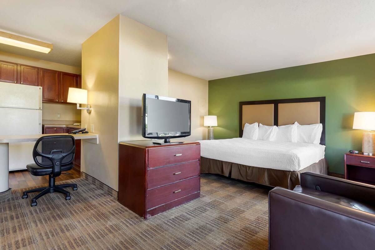 A hotel room with a bed, desk, chair, television, and a small kitchenette.