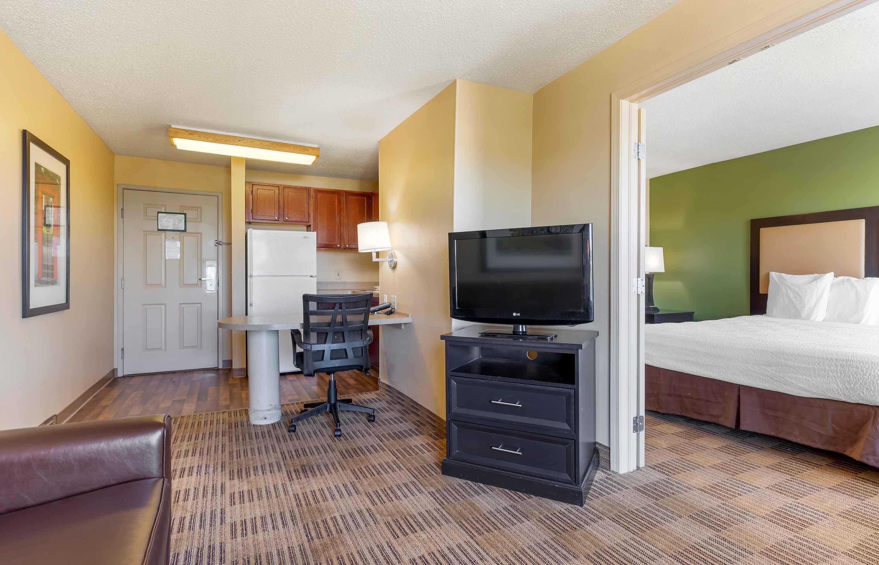 A hotel room with a bed, a TV, a desk with a chair, a refrigerator, and a small kitchenette.