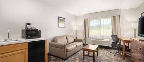 Una habitación de hotel con kitchenette, sofá, escritorio, televisión y ventana con cortinas.
