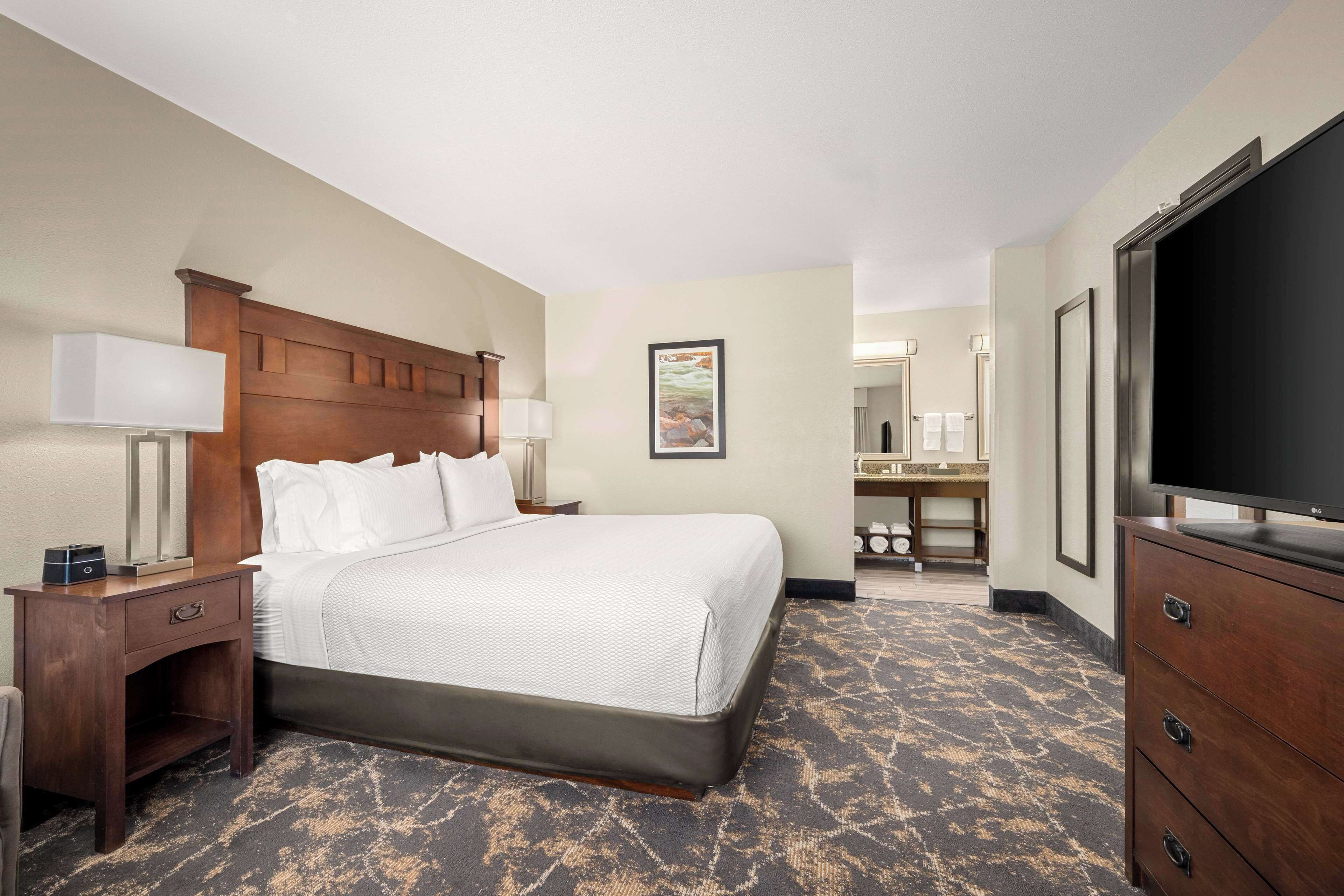 A hotel room with a large bed, a wooden headboard, a nightstand with a lamp, a dresser, a television, and a bathroom visible in the background.