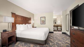A hotel room with a large bed, a wooden headboard, a nightstand with a lamp, a dresser, a television, and a bathroom visible in the background.