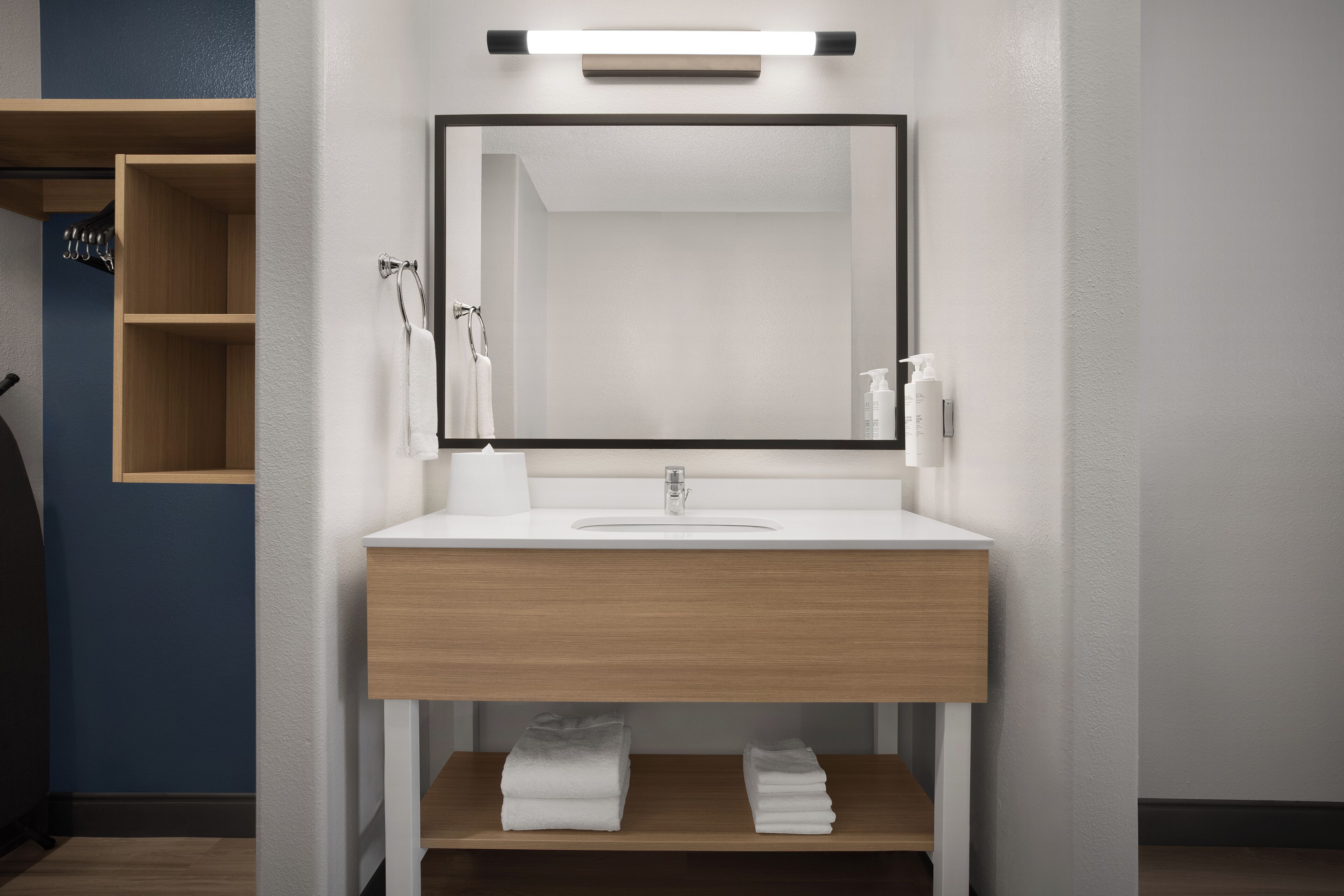 A modern bathroom with a wooden vanity, a large mirror, and a shelf with folded towels.