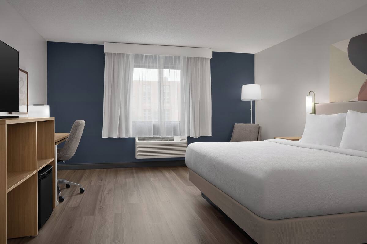 A hotel room with a bed, a desk, a chair, a TV, and a window with curtains.