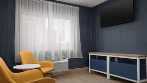 Een kamer met een groot raam, een flatscreen-tv aan de muur, een klein rond tafeltje en twee gele stoelen.
