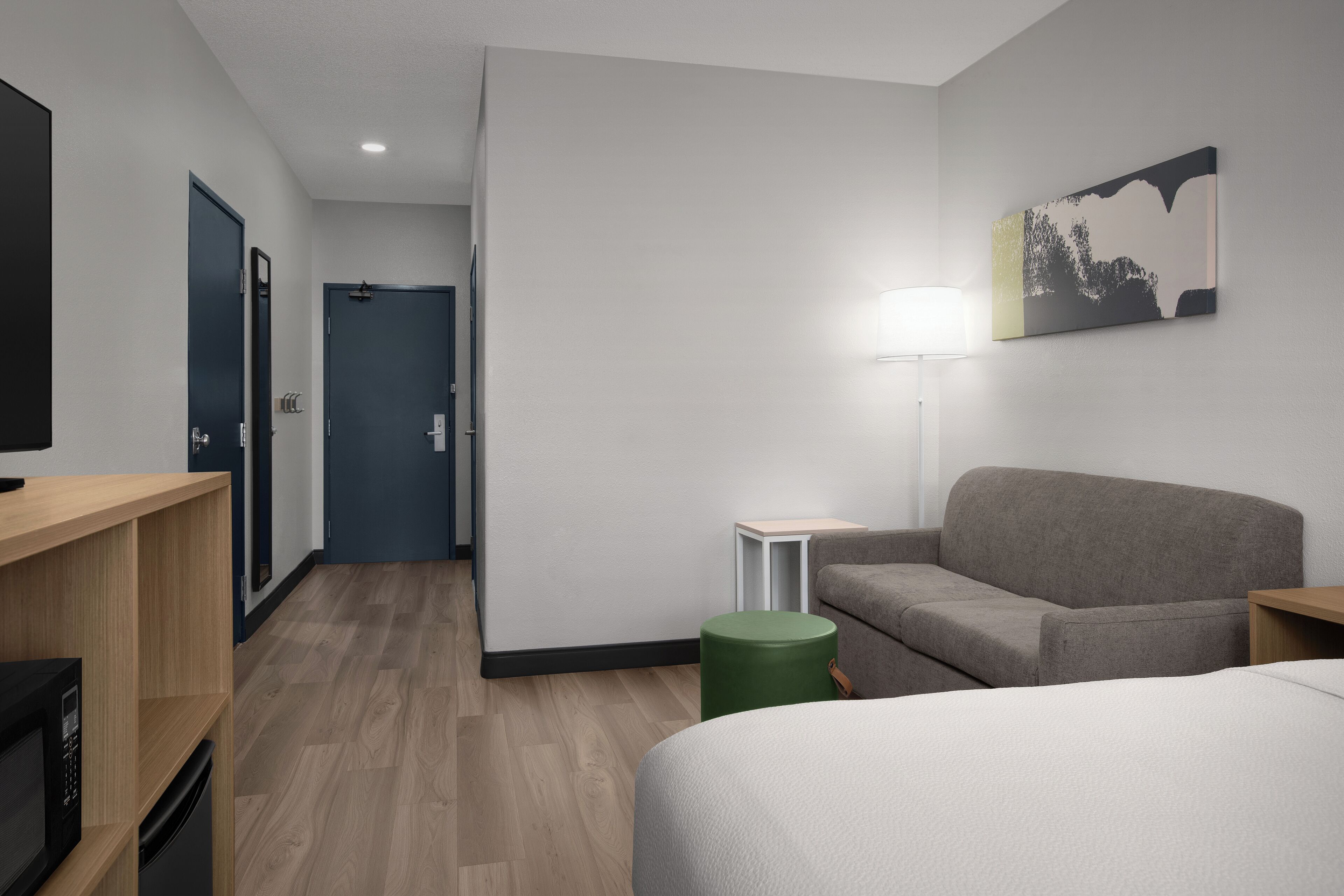 A modern hotel room with a wooden floor, a grey sofa, a green ottoman, a bed, a TV, and a painting on the wall.