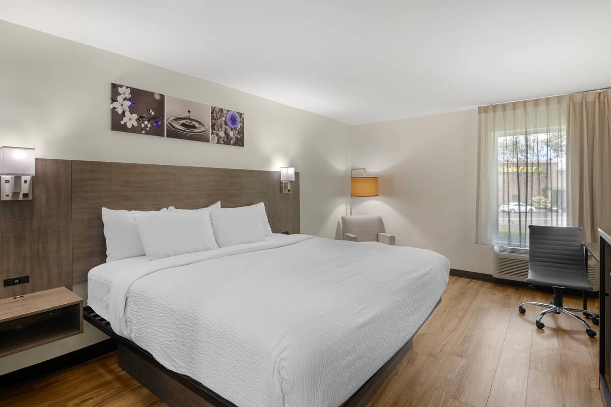 A hotel room with a large bed, a desk with a chair, a window with curtains, and wall-mounted lamps.