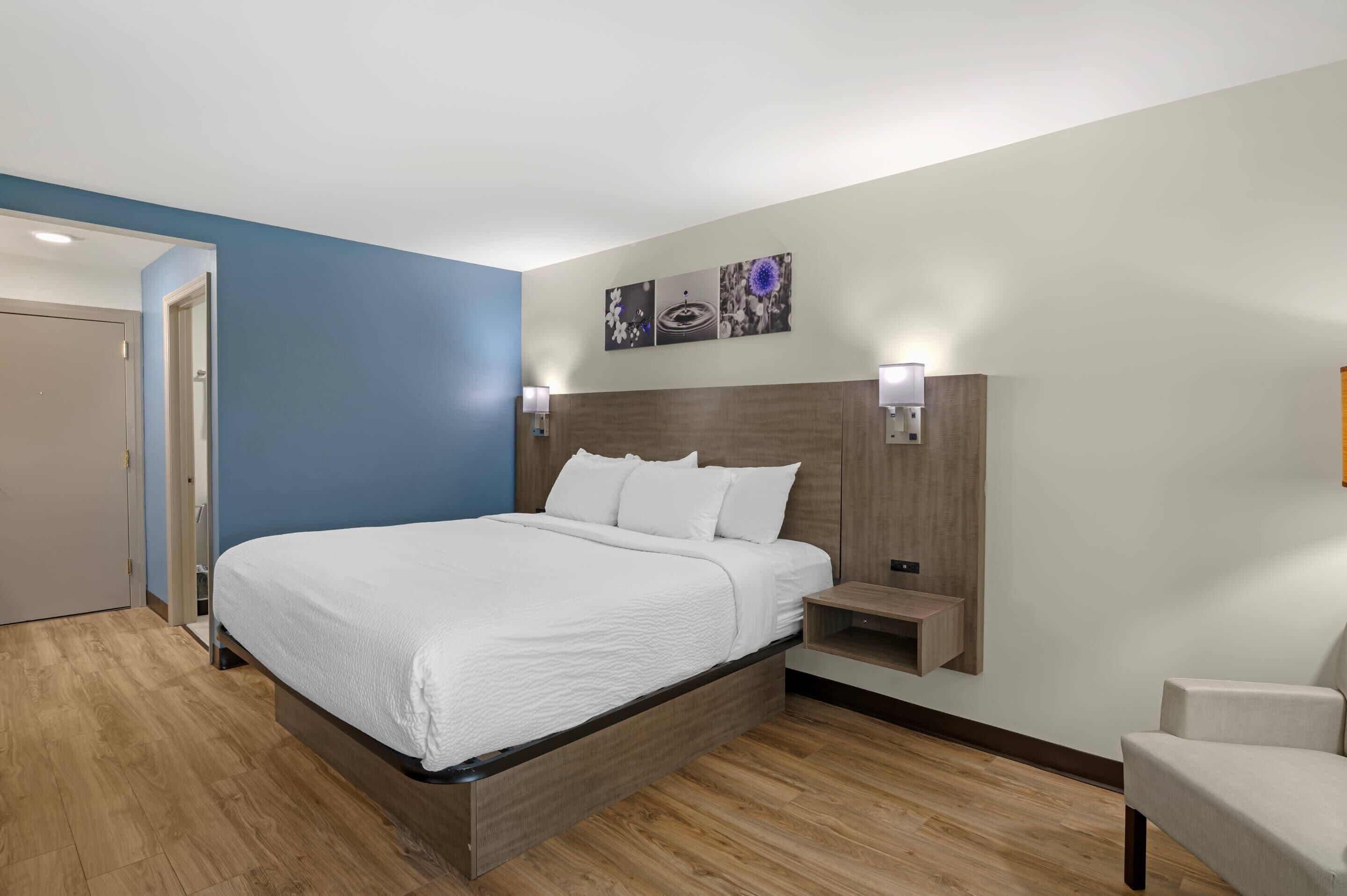 A modern hotel room with a large bed, a bedside table, a wall-mounted shelf, and a door leading to another room.