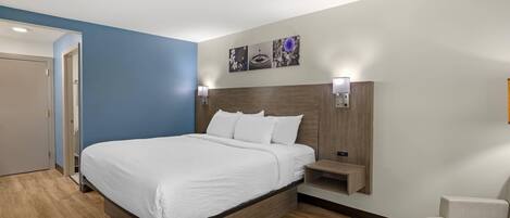A modern hotel room with a large bed, a bedside table, a wall-mounted shelf, and a door leading to another room.