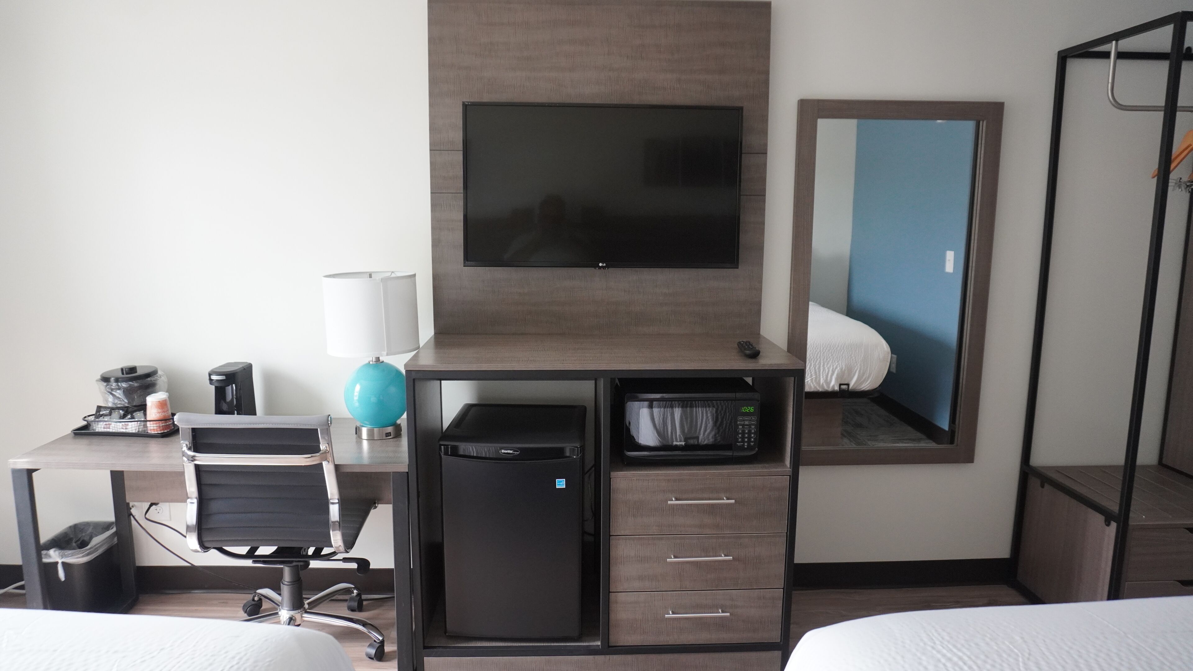 Una habitación de hotel con cama, escritorio con computadora, cafetera, lámpara, televisor y espejo.