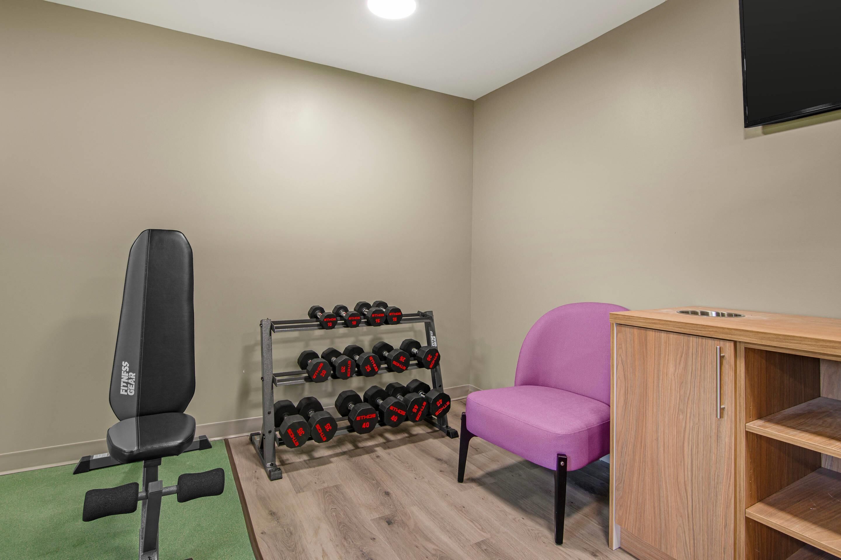 A gym with a bench, dumbbells, a pink chair, and a wooden cabinet.