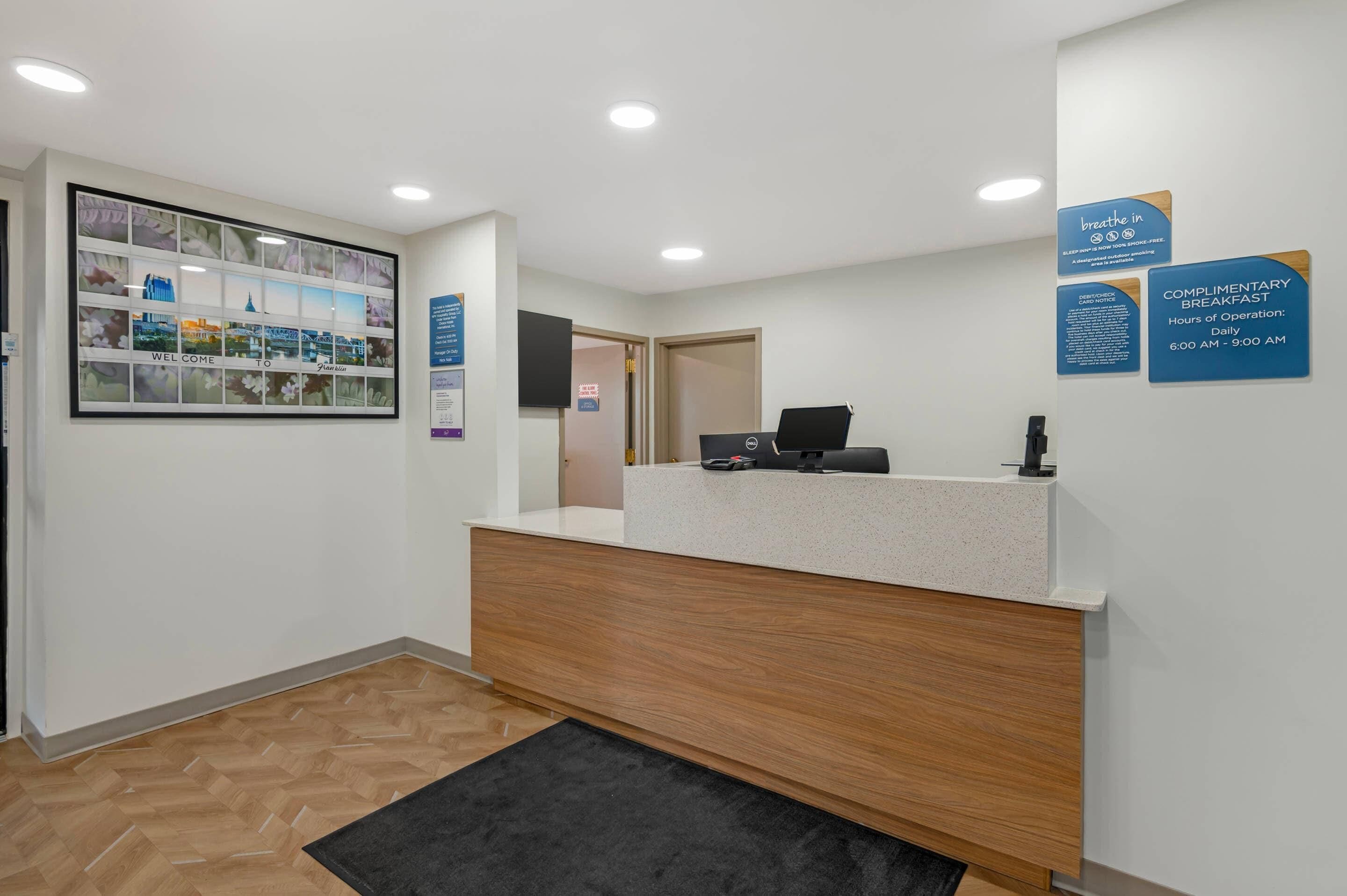 A reception desk with a computer, a 'Complimentary Breakfast' sign, and a 'Welcome' photo wall.