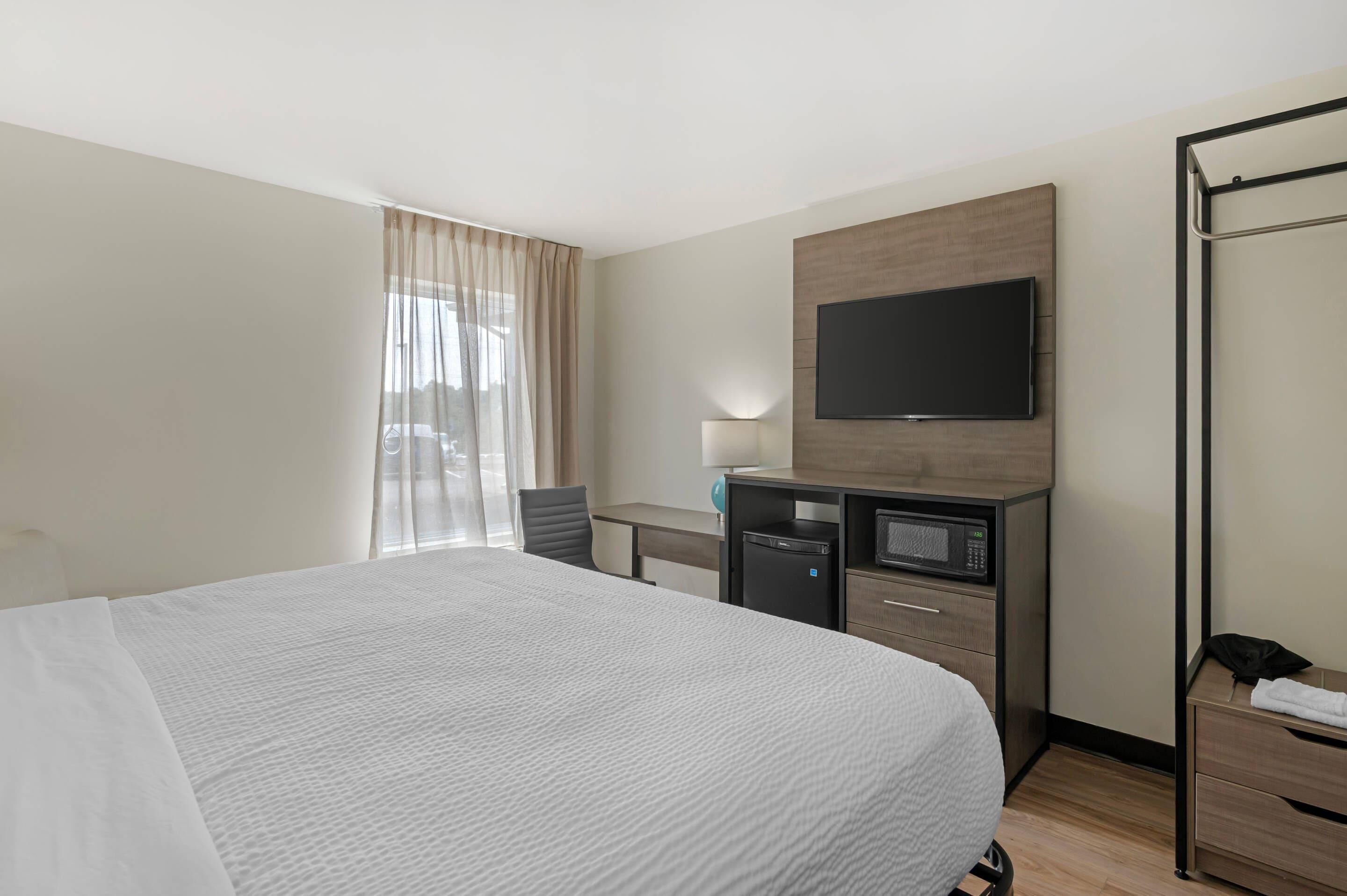A hotel room with a large bed, a desk with a computer, a TV mounted on the wall, and a window with sheer curtains.