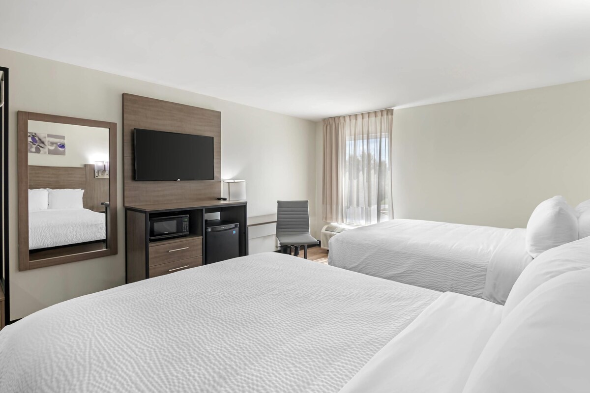 Quarto de hotel com duas camas, televisão, escrivaninha com cadeira e janela com cortinas.