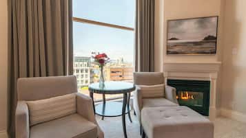 Habitación de hotel con dos sillones, una mesita redonda con un florero, chimenea y vistas a la ciudad a través de amplias ventanas.