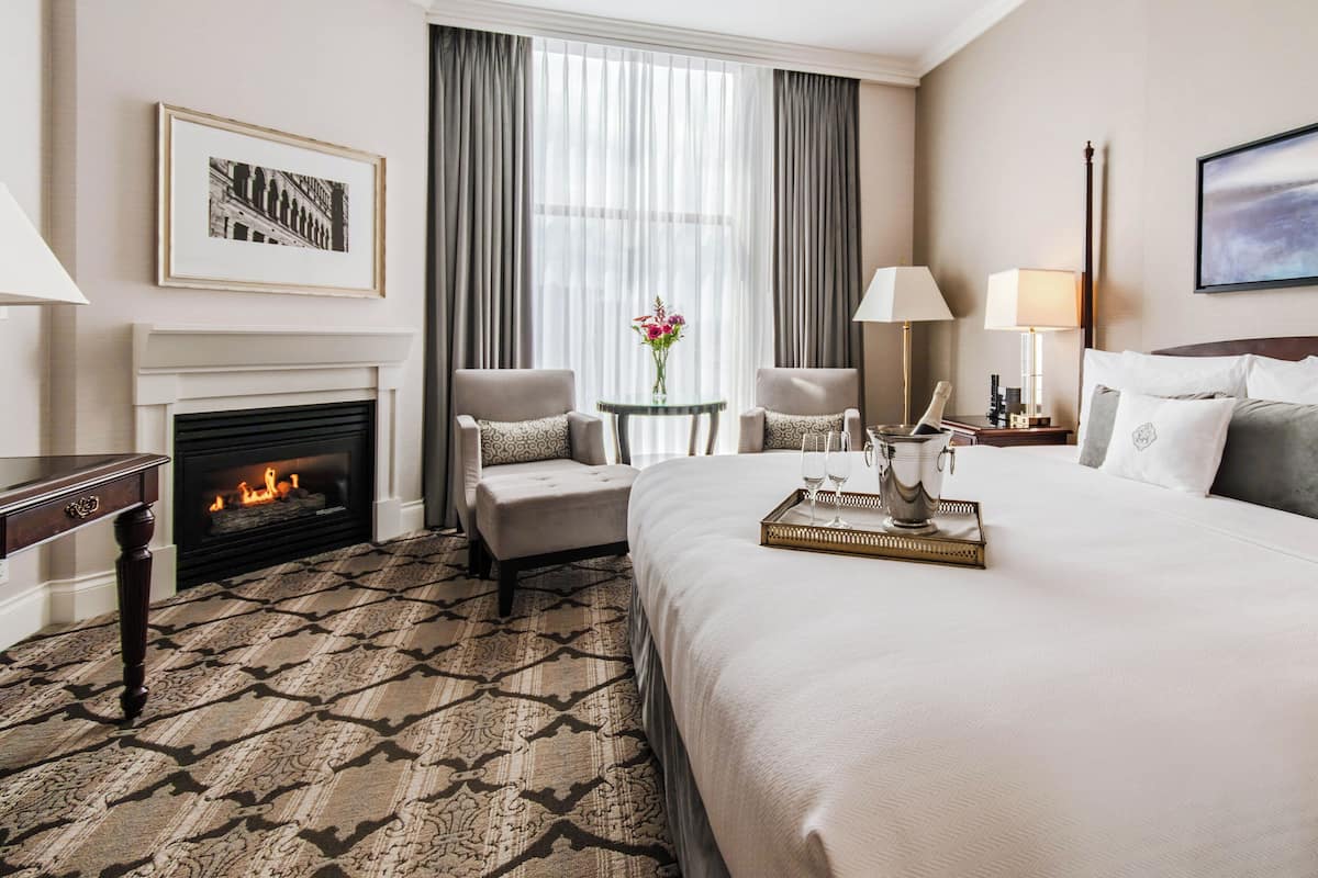 Habitación de hotel con una cama grande, una chimenea, un rincón de estar con dos sillones, una mesita con un jarrón de flores y una mesita de noche con una lámpara.