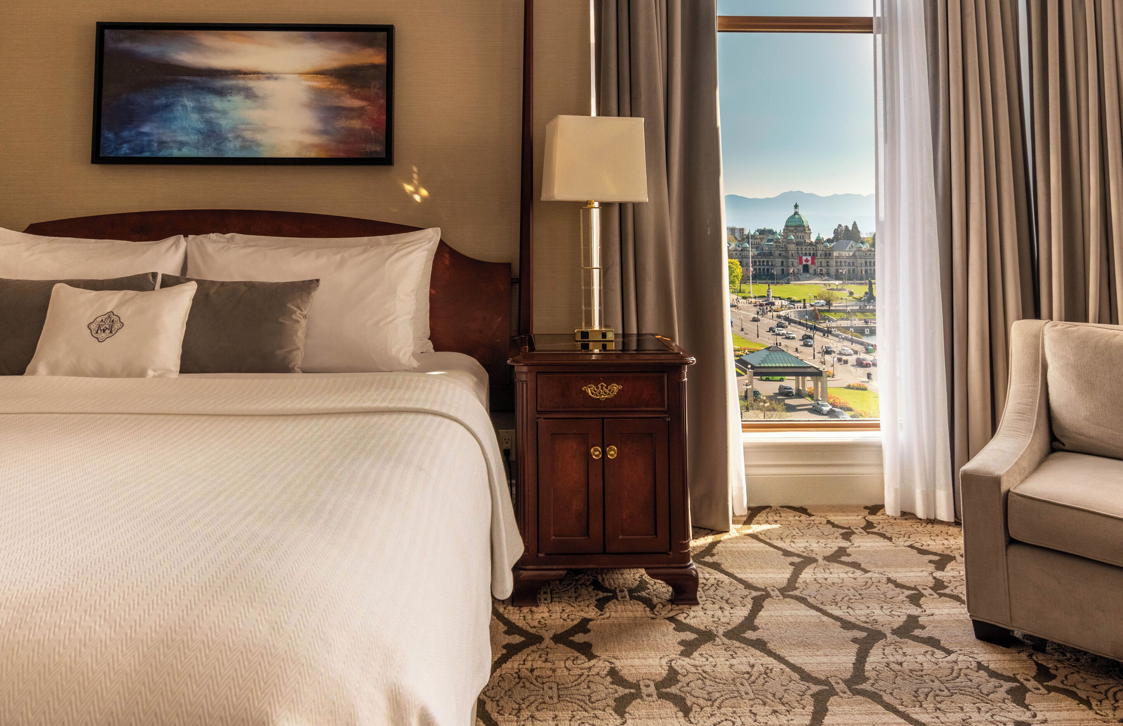 A hotel room with a large bed, a nightstand with a lamp, a view of a cityscape through a window, and a chair.
