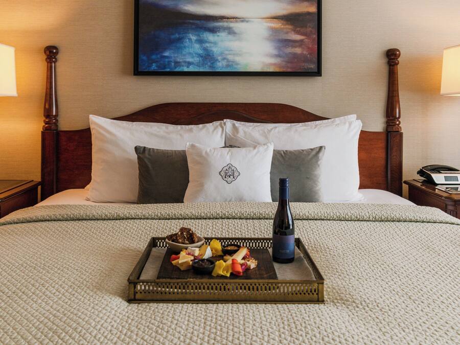 A neatly made bed with a tray of fruit and a bottle of wine, a painting on the wall, and two bedside lamps.
