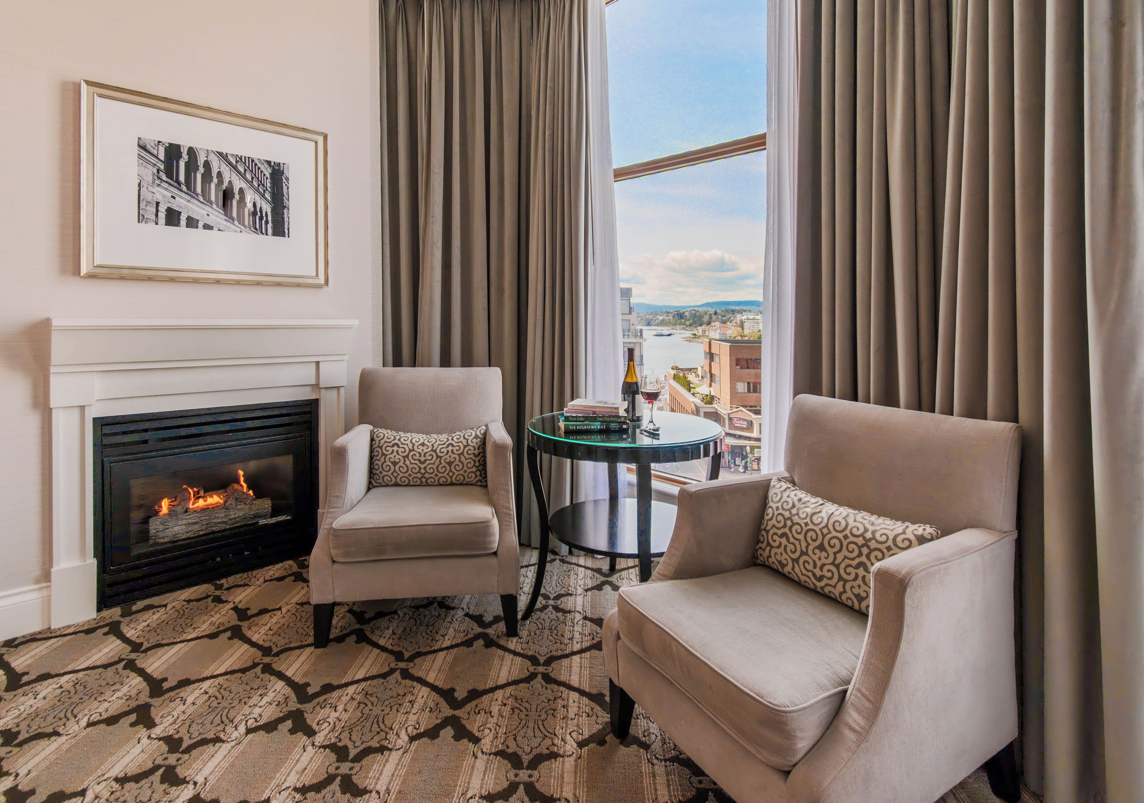 A room with a fireplace, two armchairs, a small table with books, and a view of a cityscape through large windows.