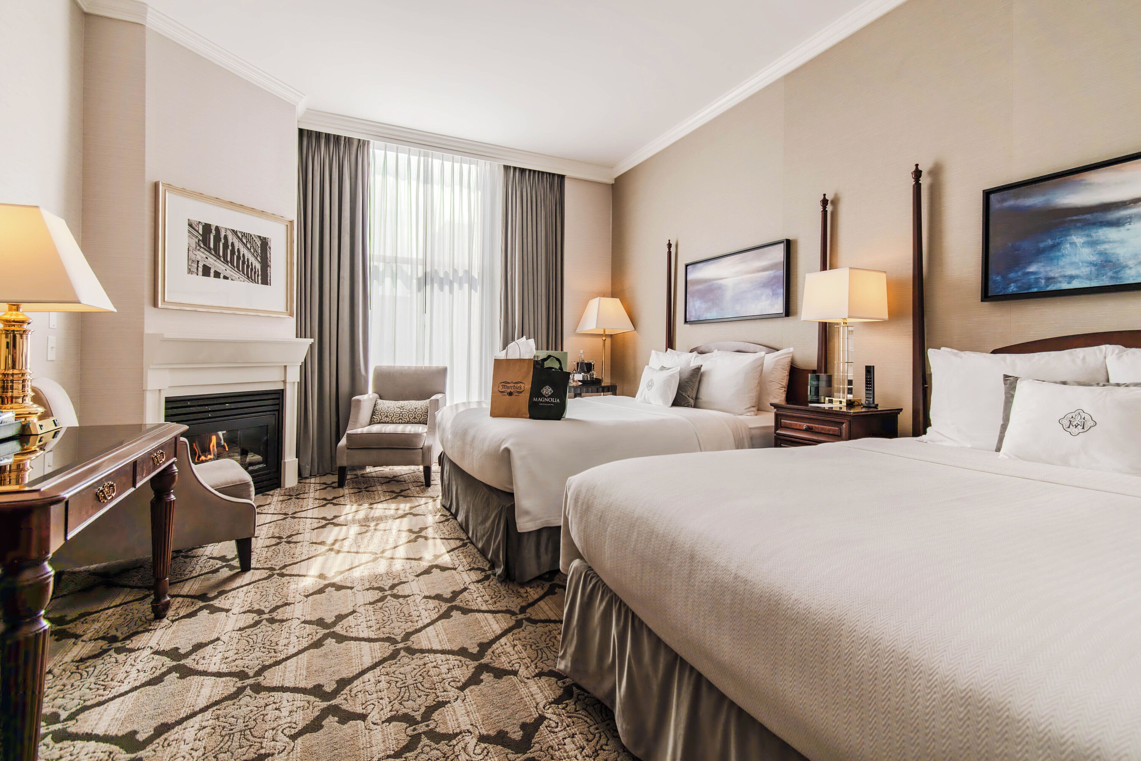 A hotel room with two beds, a fireplace, a desk with a lamp, and a chair.