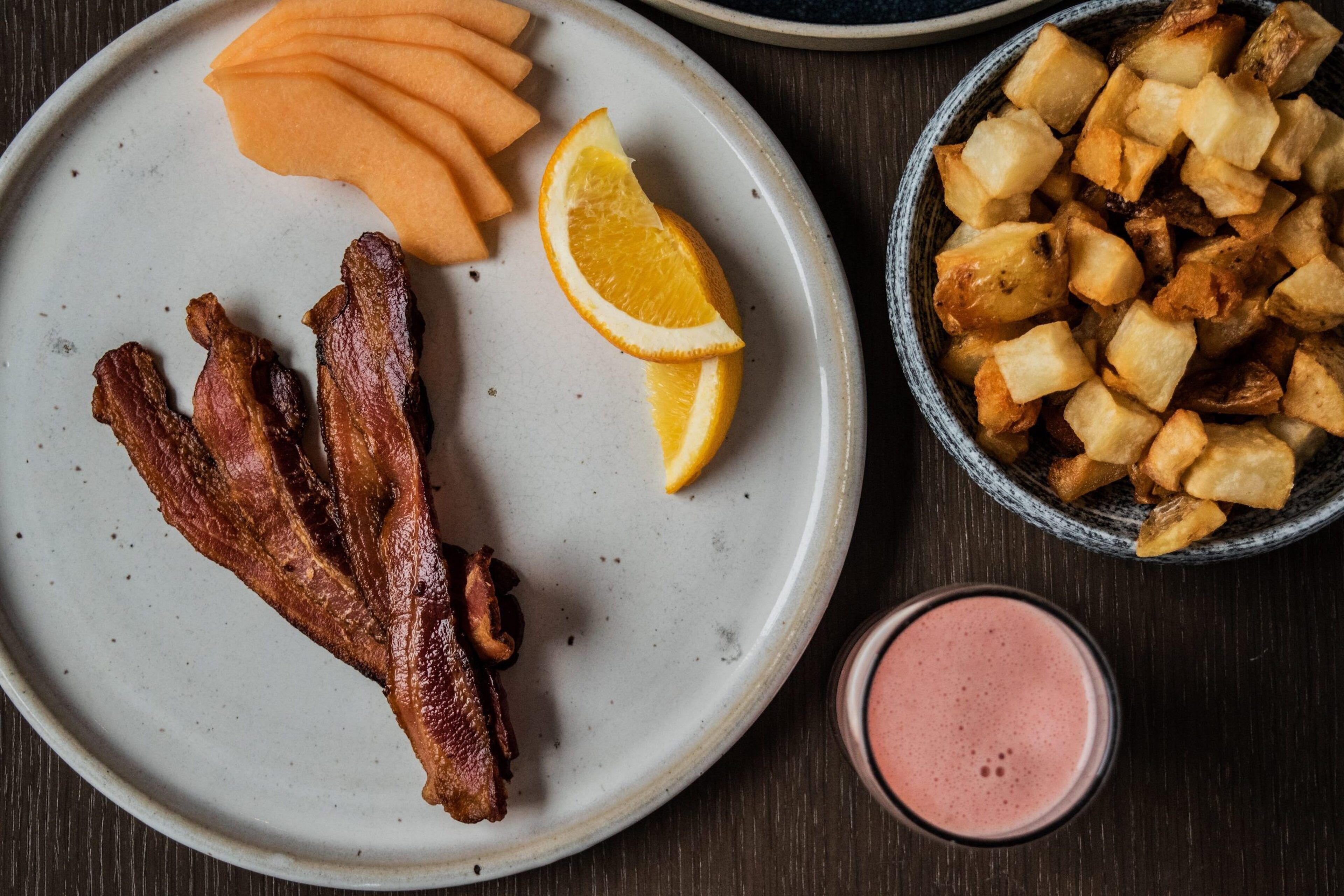 Un plato con tocino, rodajas de naranja y un bol de patatas.
