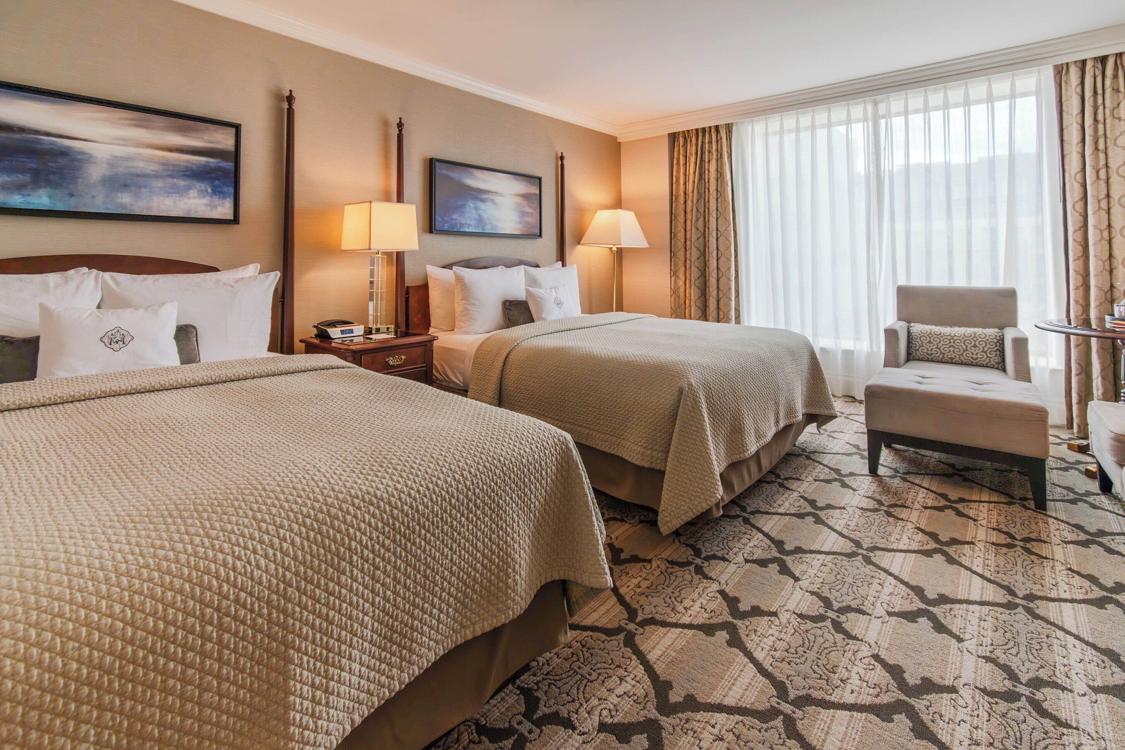A hotel room with two beds, a chair, a desk, and a large window with curtains.