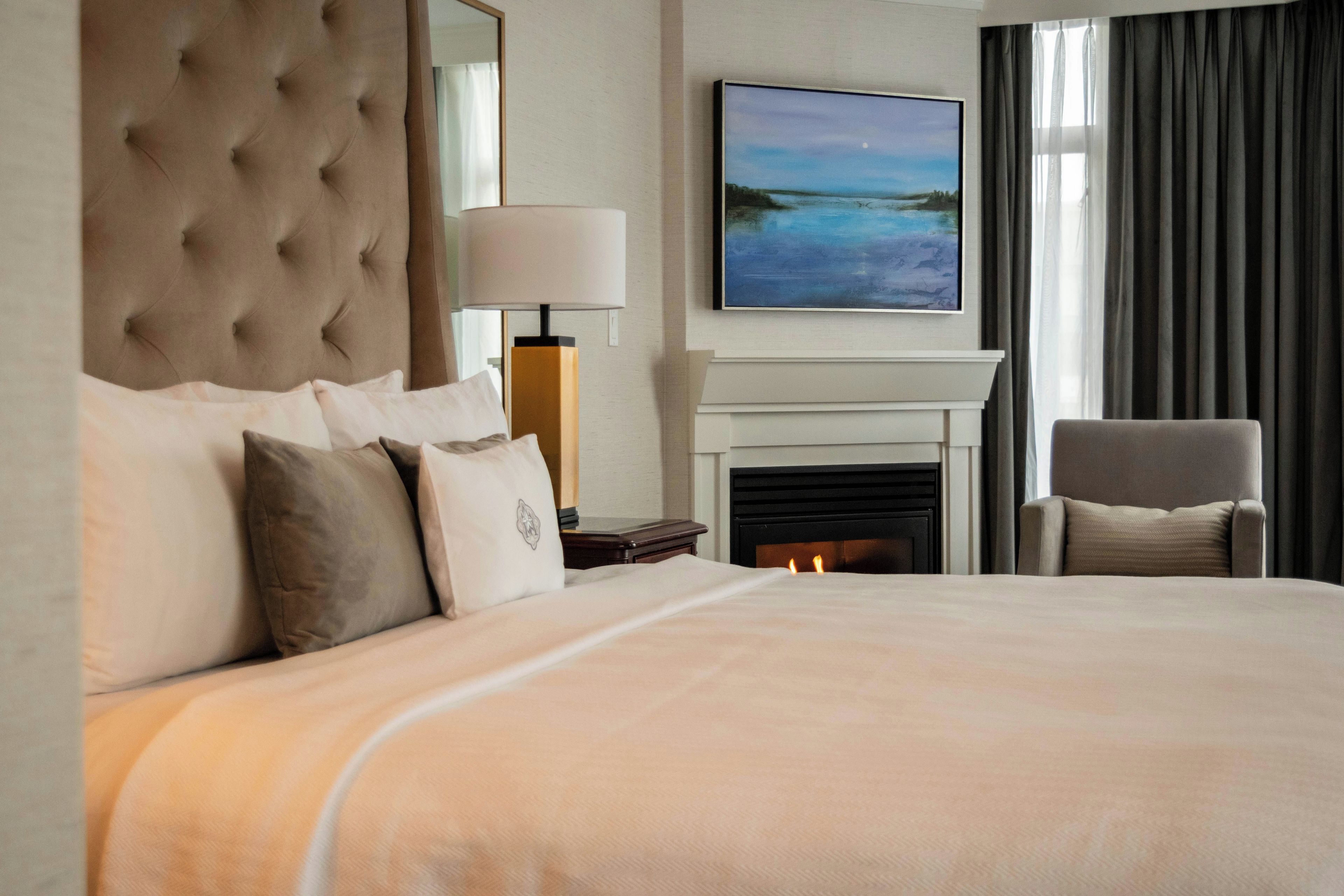 A hotel room with a large bed, a fireplace, a nightstand, a lamp, a chair, and a painting on the wall.