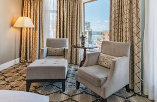 Habitación de hotel con dos sillones, una mesita con una botella y vasos, y vista a un paisaje urbano a través de amplios ventanales.
