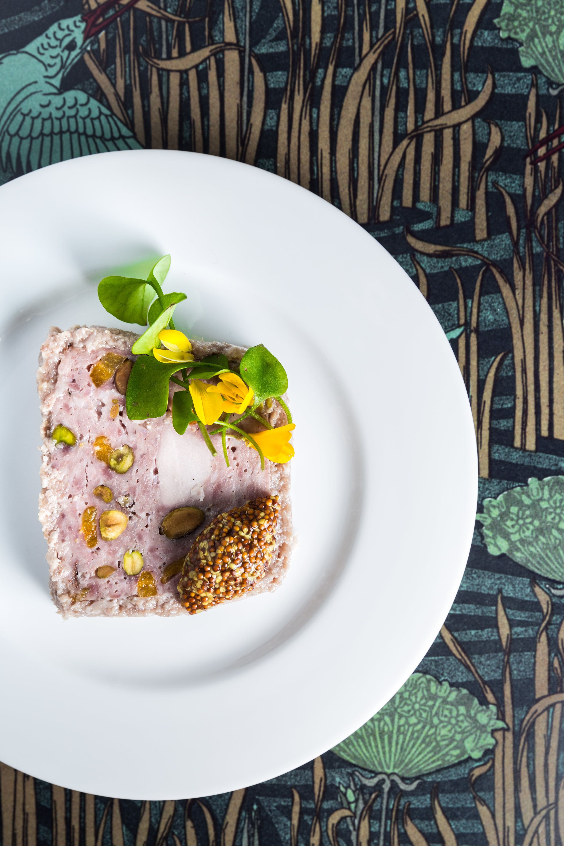 A slice of meatloaf with nuts and seeds on a white plate, set against a patterned tablecloth.