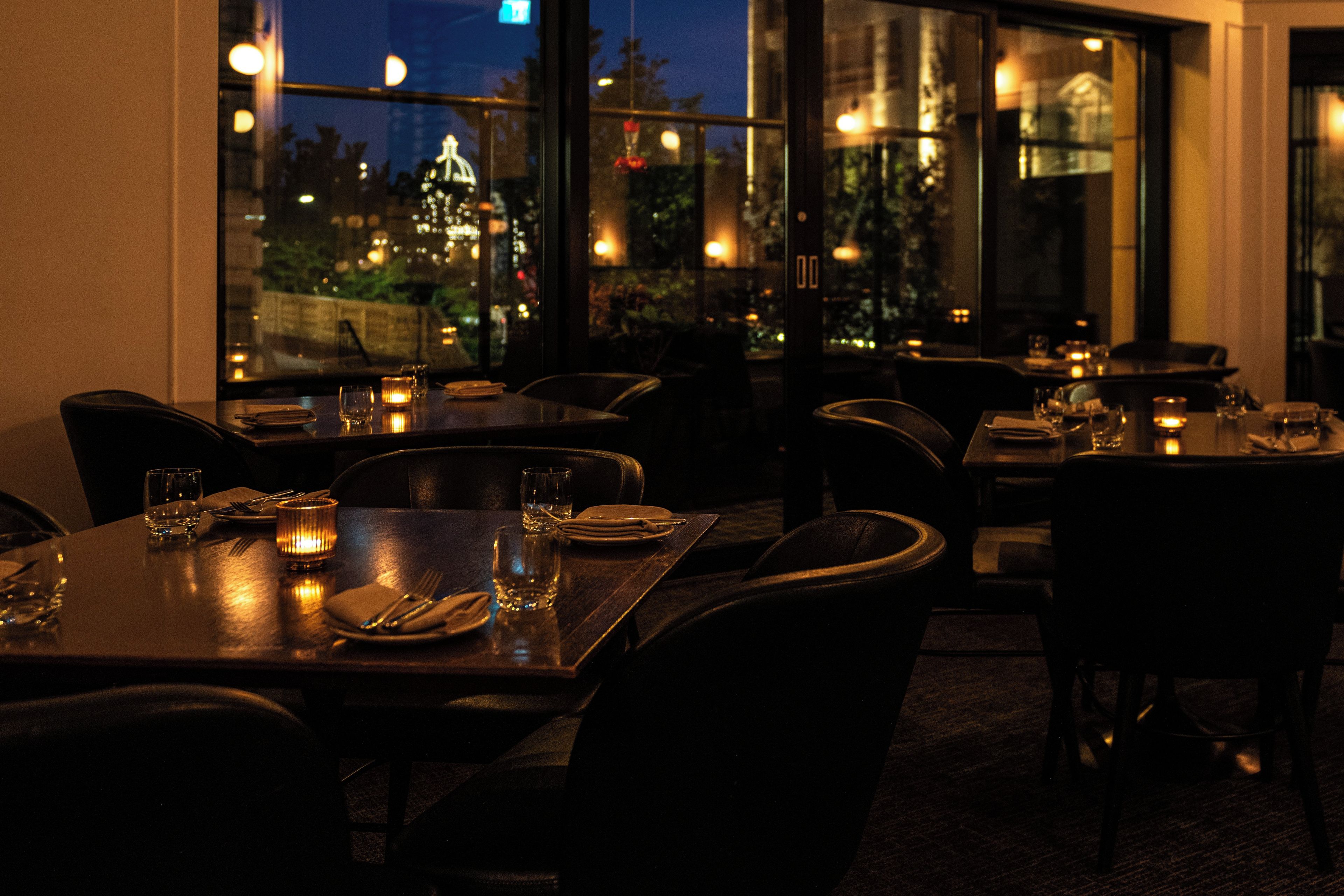 A dimly lit restaurant with tables set for dining, large windows, and a view of the cityscape at night.