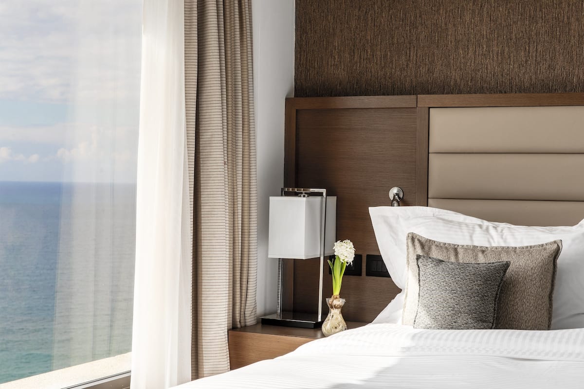 Una habitación de hotel con una cama, una mesita de noche con lámpara, un jarrón con flores y vista al océano.