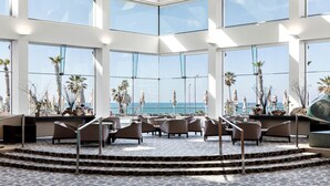 Un vaste espace intérieur doté de grandes fenêtres offrant une vue sur la mer. On y trouve plusieurs coins salon avec des chaises et des tables.