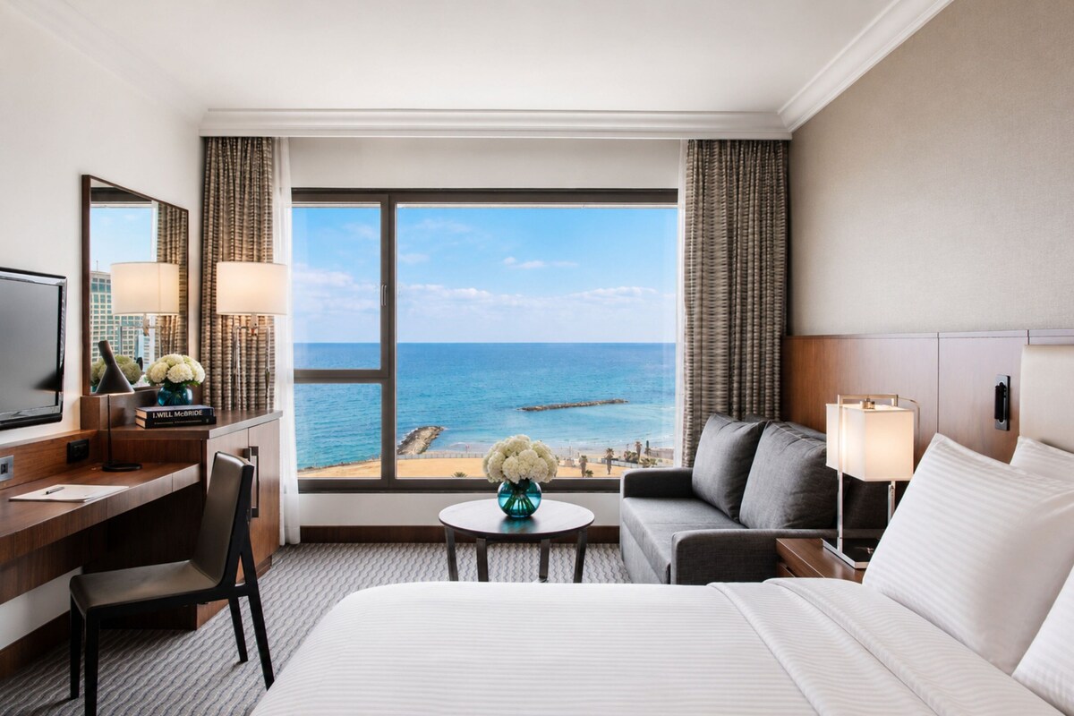 A hotel room with a bed, a desk, a chair, a sofa, a small table with a vase, and a large window offering a view of the sea.