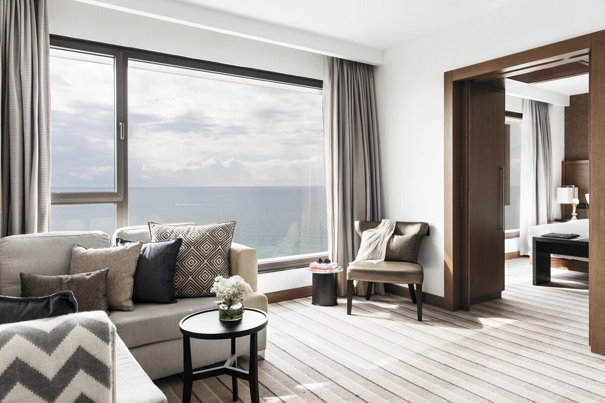 A modern hotel room with a large window offering a sea view, a cozy seating area with a sofa and armchair, and a small round table with a vase of flowers.