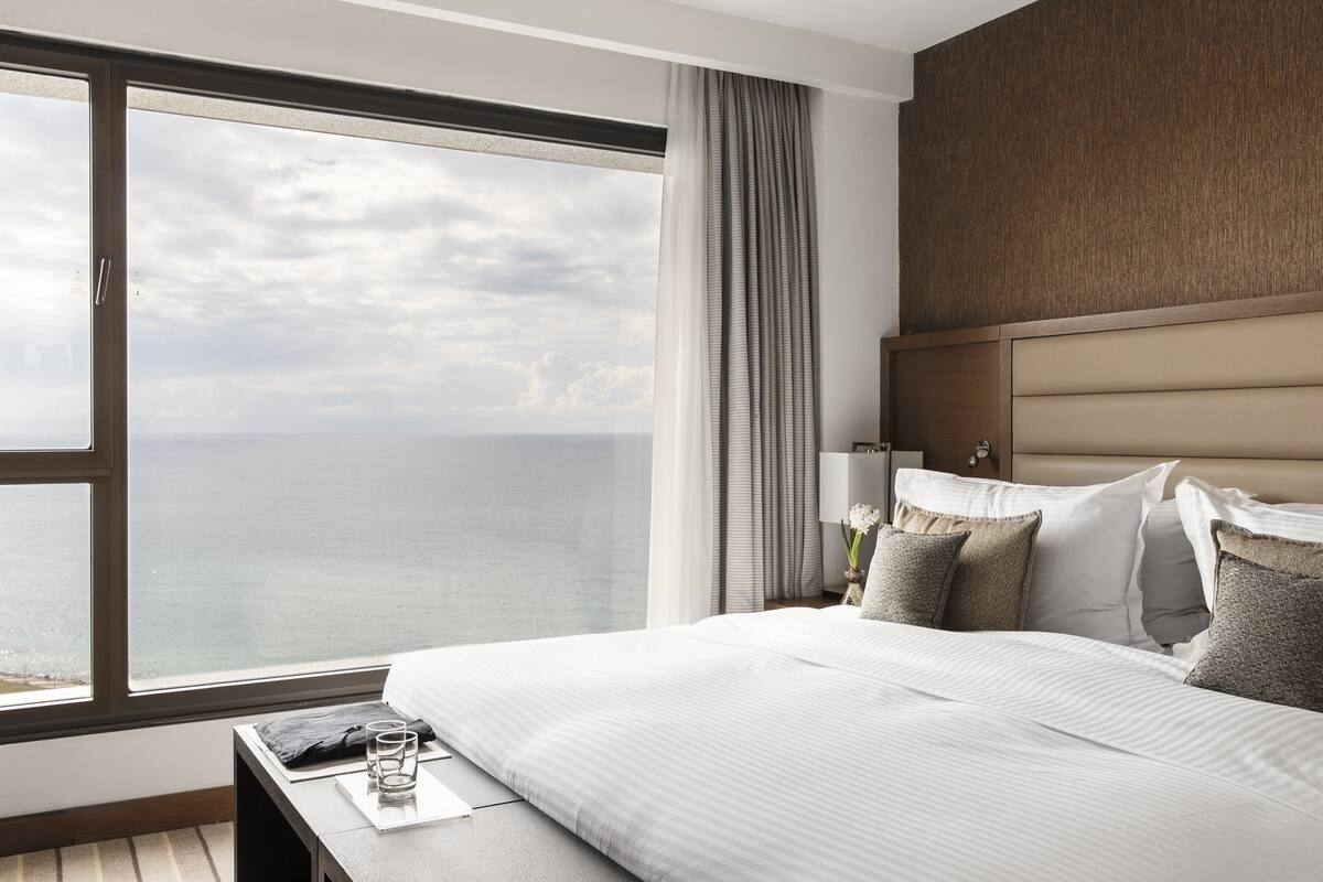 A hotel room with a large window offering a view of the sea, a bed with white bedding, a nightstand with a lamp, and a wooden headboard.