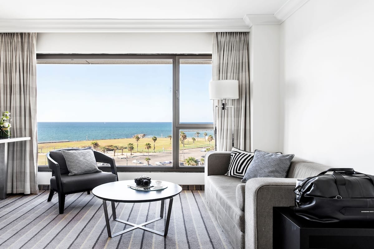 Habitación de hotel con un ventanal grande que ofrece vista a la playa, un sofá, una silla, una mesita, un televisor y una maleta.