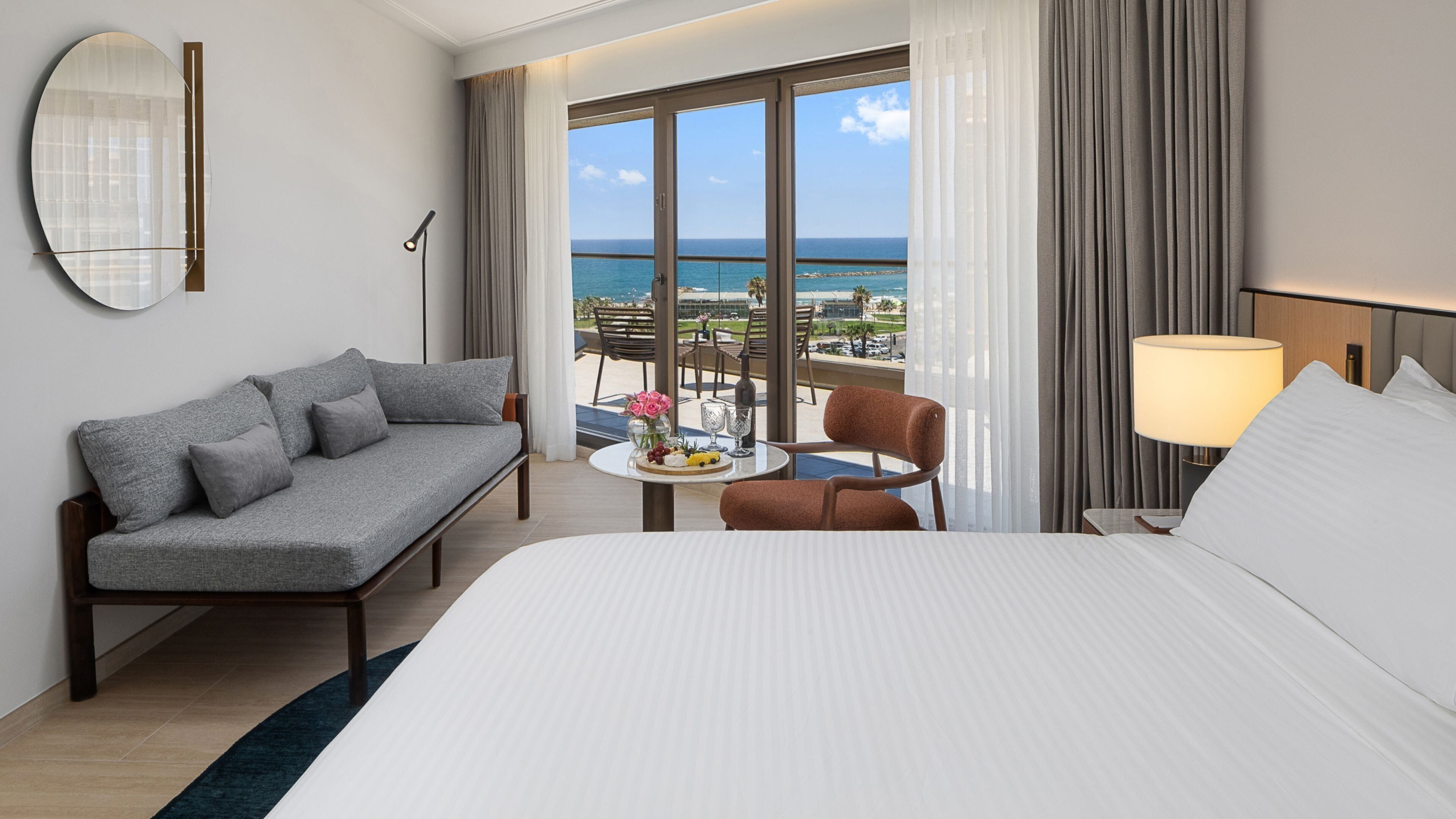A hotel room with a bed, a sofa, a chair, a small table with a fruit platter, and a view of the ocean.