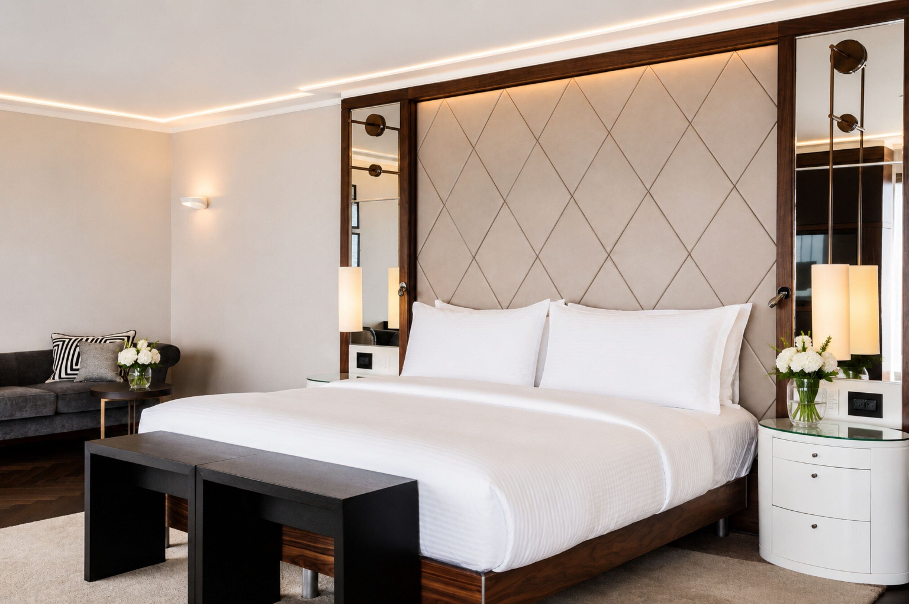Un dormitorio moderno con una cama grande, mesitas de noche, un taburete y un espejo decorativo.