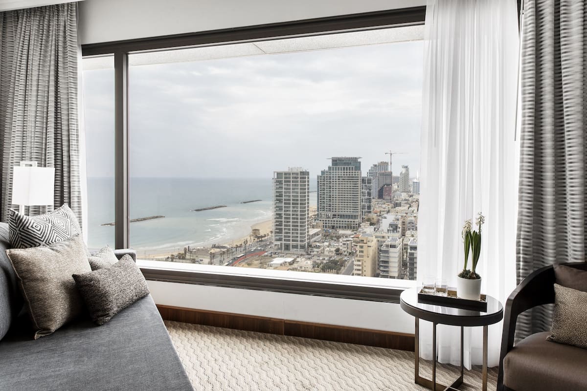 Una habitación de hotel moderna con un ventanal amplio que ofrece vistas a la ciudad, un sofá con cojines estampados, una mesita redonda con una planta en maceta y una silla con cojín.