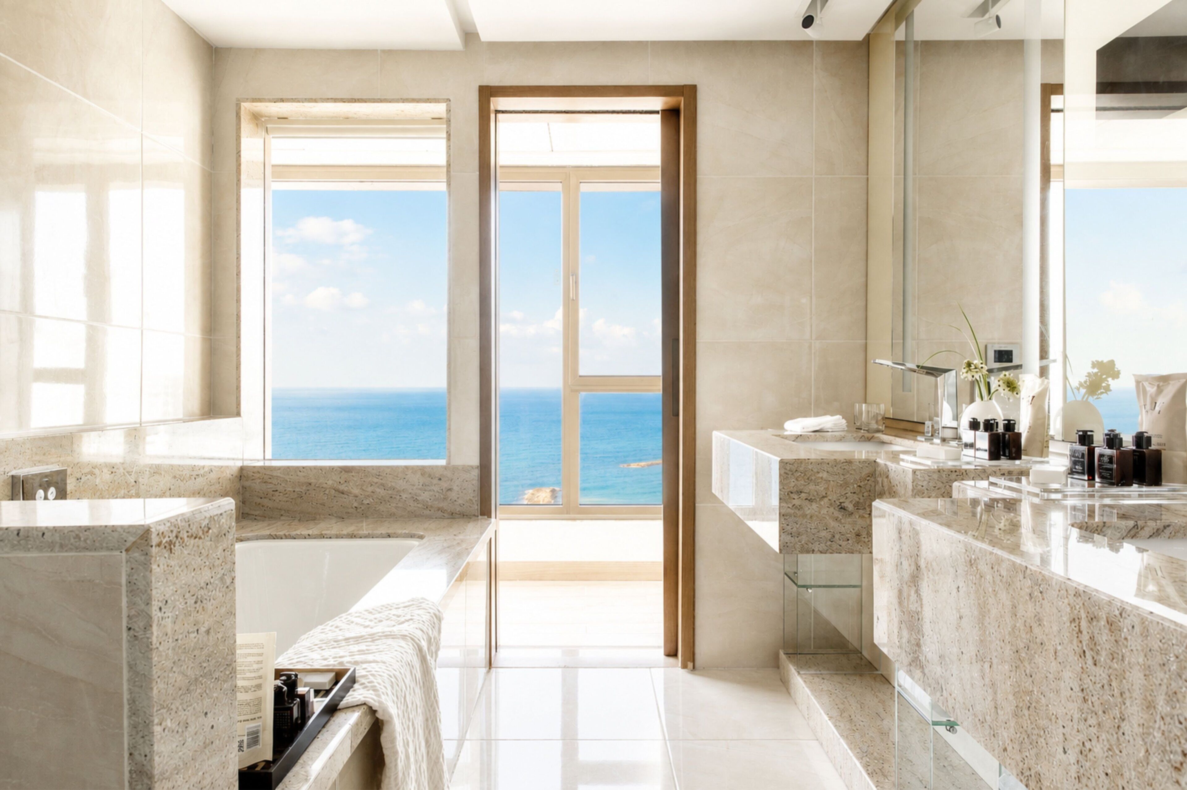 A modern bathroom with a large window offering a view of the sea, a marble sink, and a glass shower enclosure.