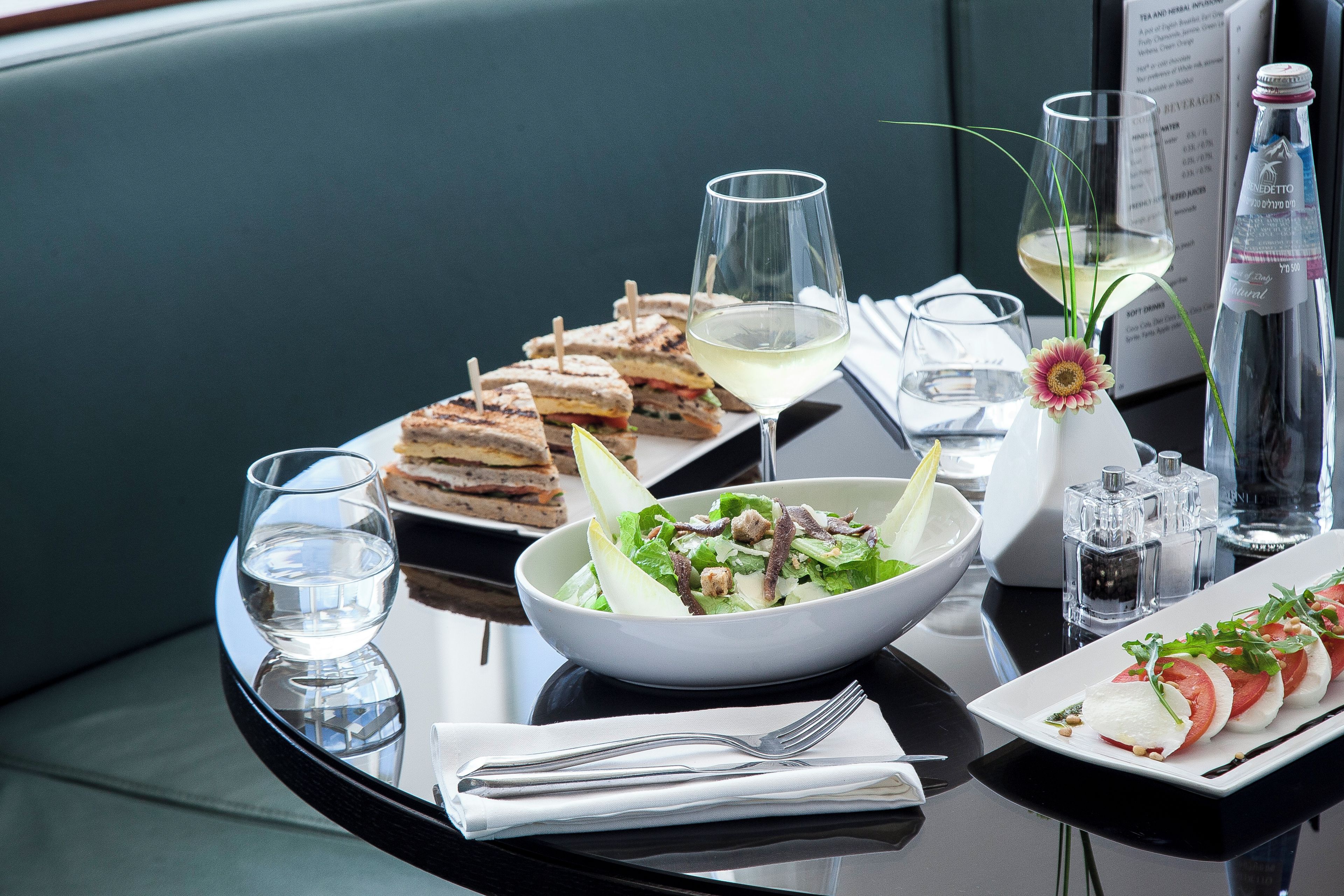 A dining table with a variety of food items, including sandwiches, a salad, and a plate of sliced tomatoes, accompanied by glasses and a bottle of water.