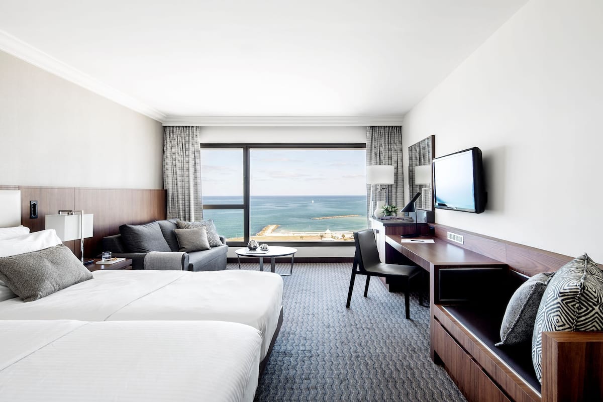 A hotel room with two beds, a desk, a chair, a TV, and a large window with a view of the sea.