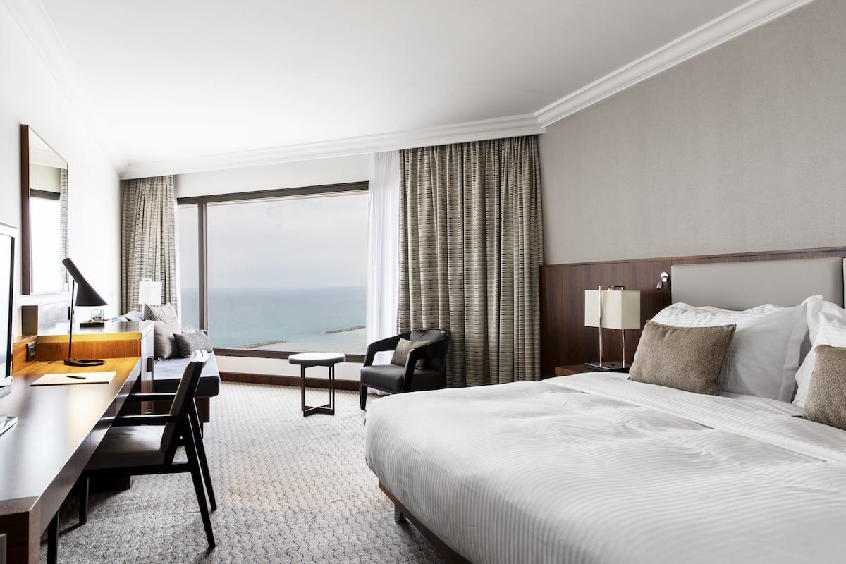 A hotel room with a large bed, a desk with a chair, a small table, and a view of the ocean.
