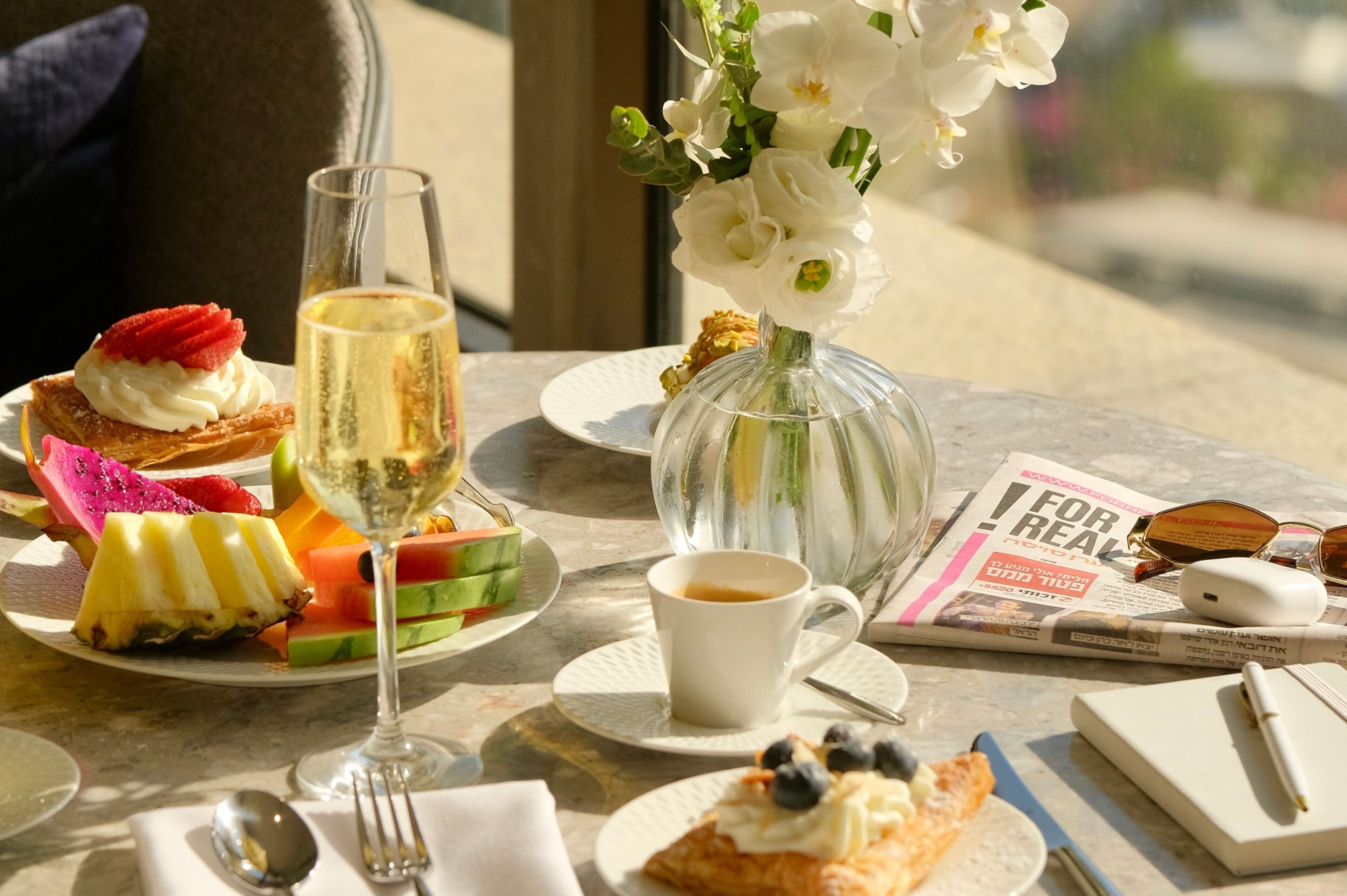 Una mesa con diversas opciones para el desayuno, una copa de champán, un jarrón de flores y una revista.