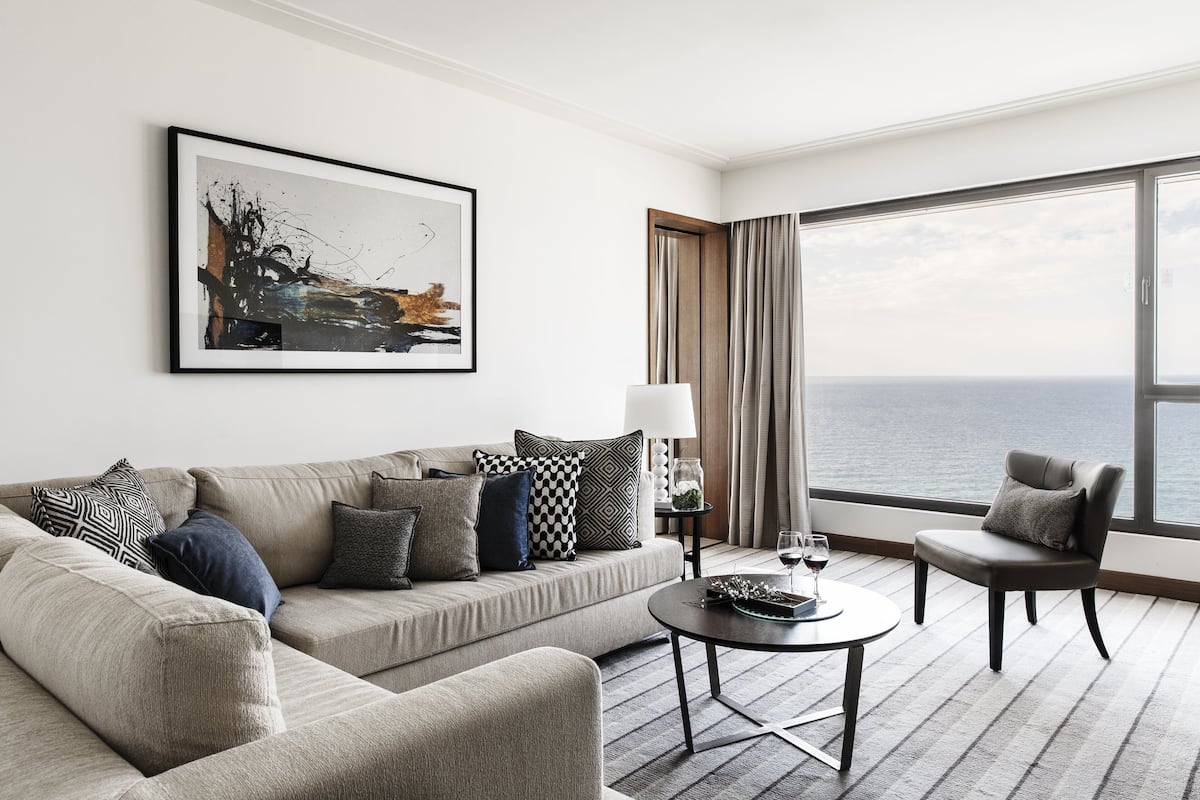 Un hotel moderno con un ventanal grande que ofrece vistas al mar, un sofá cómodo con cojines estampados, una pequeña mesa redonda con una copa de vino y un cuadro abstracto enmarcado en la pared.