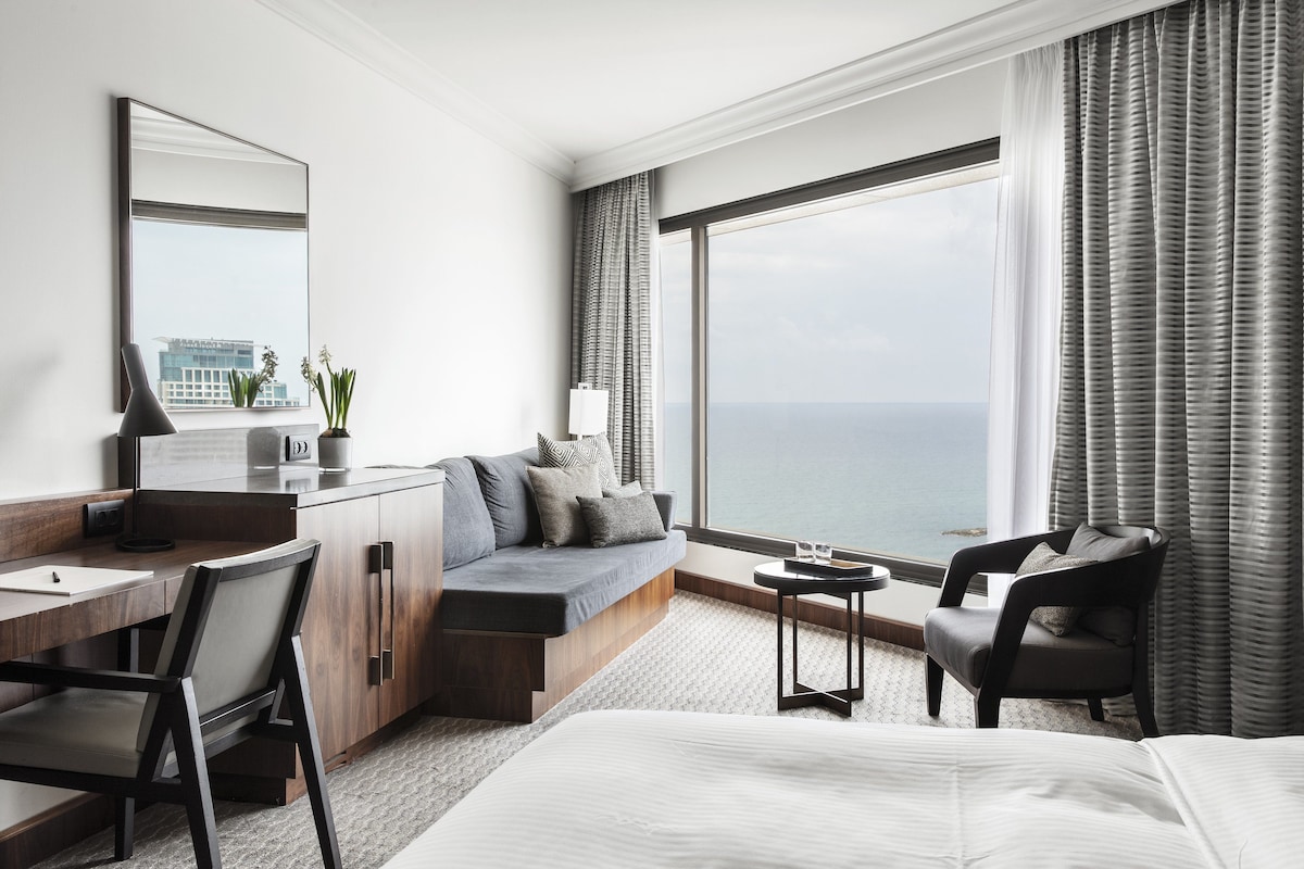 Ein modernes Hotelzimmer mit großem Fenster, einer Couch, einem Schreibtisch, einem Stuhl und einem Bett.