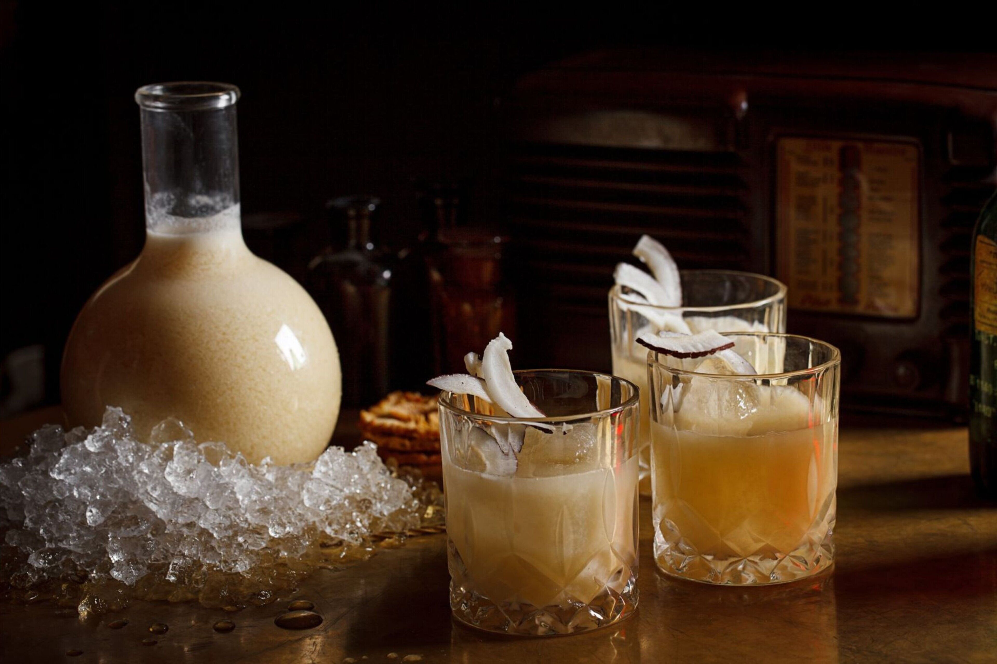 A bottle and two glasses of a beverage with ice and a garnish, placed on a table with a dark background and vintage decor.